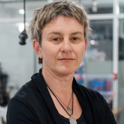 A person with short, textured hair and a neutral expression stands indoors, wearing a black top and two necklaces. The background shows a modern, glass-walled workspace.