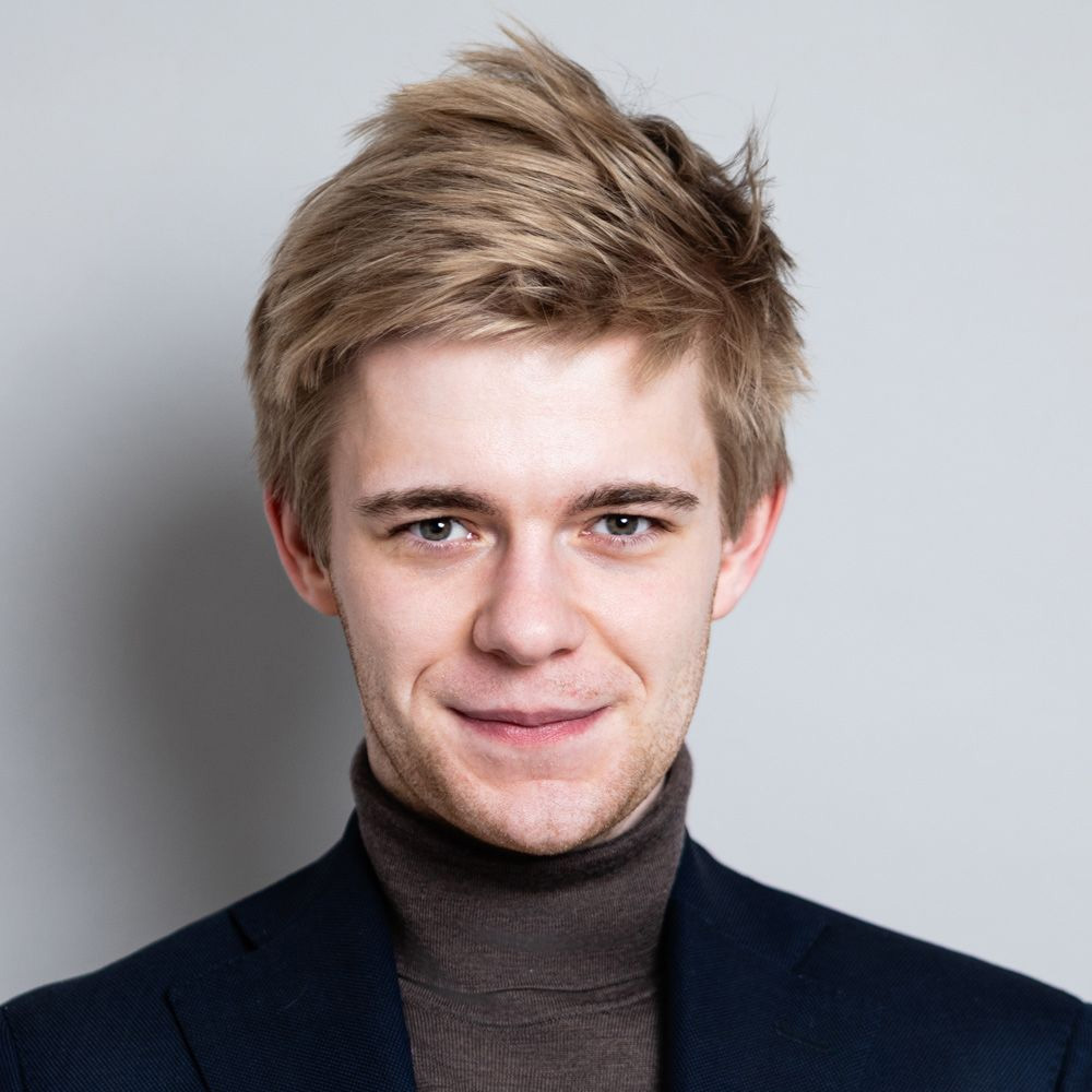 A young man with light brown hair wearing a dark turtleneck sweater and blazer, smiling in front of a plain light gray background.