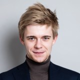 A young man with light brown hair wearing a dark turtleneck sweater and blazer, smiling in front of a plain light gray background.