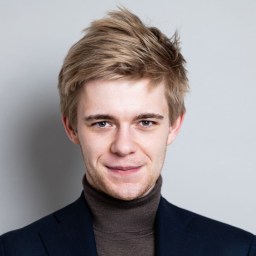 A young man with light brown hair wearing a dark turtleneck sweater and blazer, smiling in front of a plain light gray background.