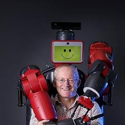 A man smiles while standing in front of a red and black robot with a screen displaying a smiley face.