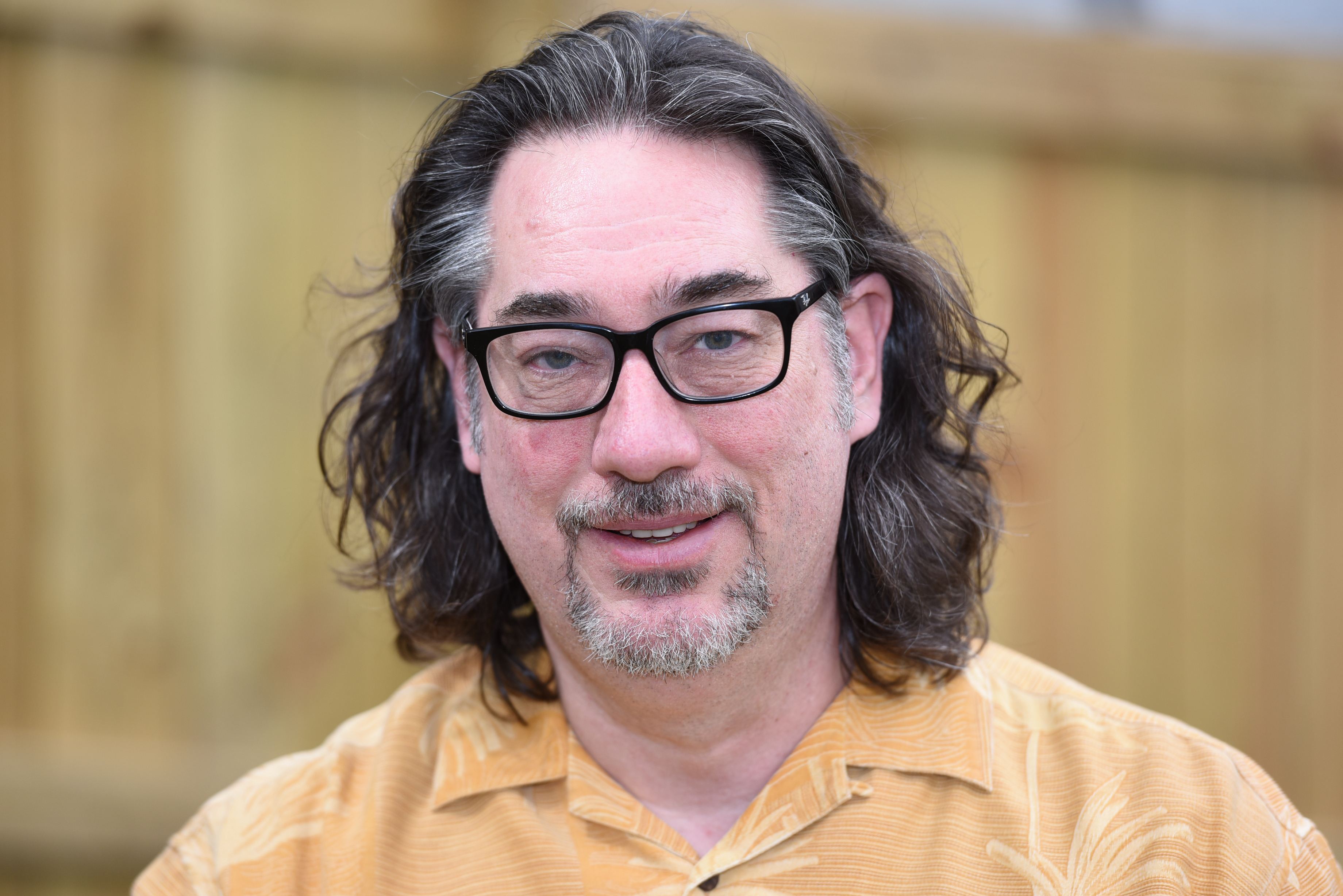 A middle-aged man with long wavy brown hair, glasses, and a short beard wearing a light brown patterned shirt stands in front of a wooden fence.