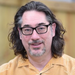 A middle-aged man with long wavy brown hair, glasses, and a short beard wearing a light brown patterned shirt stands in front of a wooden fence.