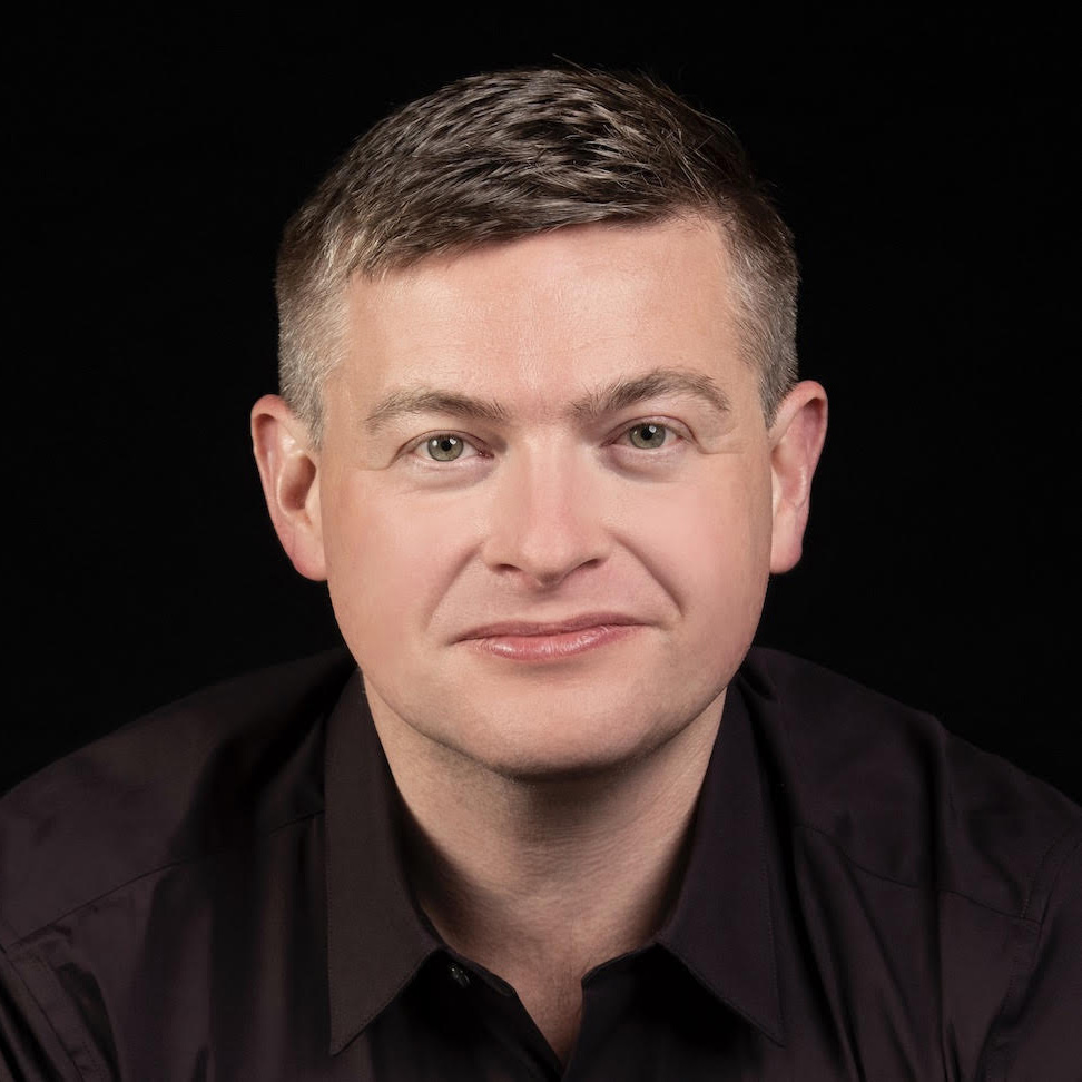 A person with short, light brown hair and light skin, wearing a dark collared shirt, is facing the camera against a black background.