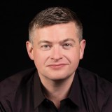 A person with short, light brown hair and light skin, wearing a dark collared shirt, is facing the camera against a black background.