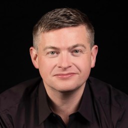 A person with short, light brown hair and light skin, wearing a dark collared shirt, is facing the camera against a black background.