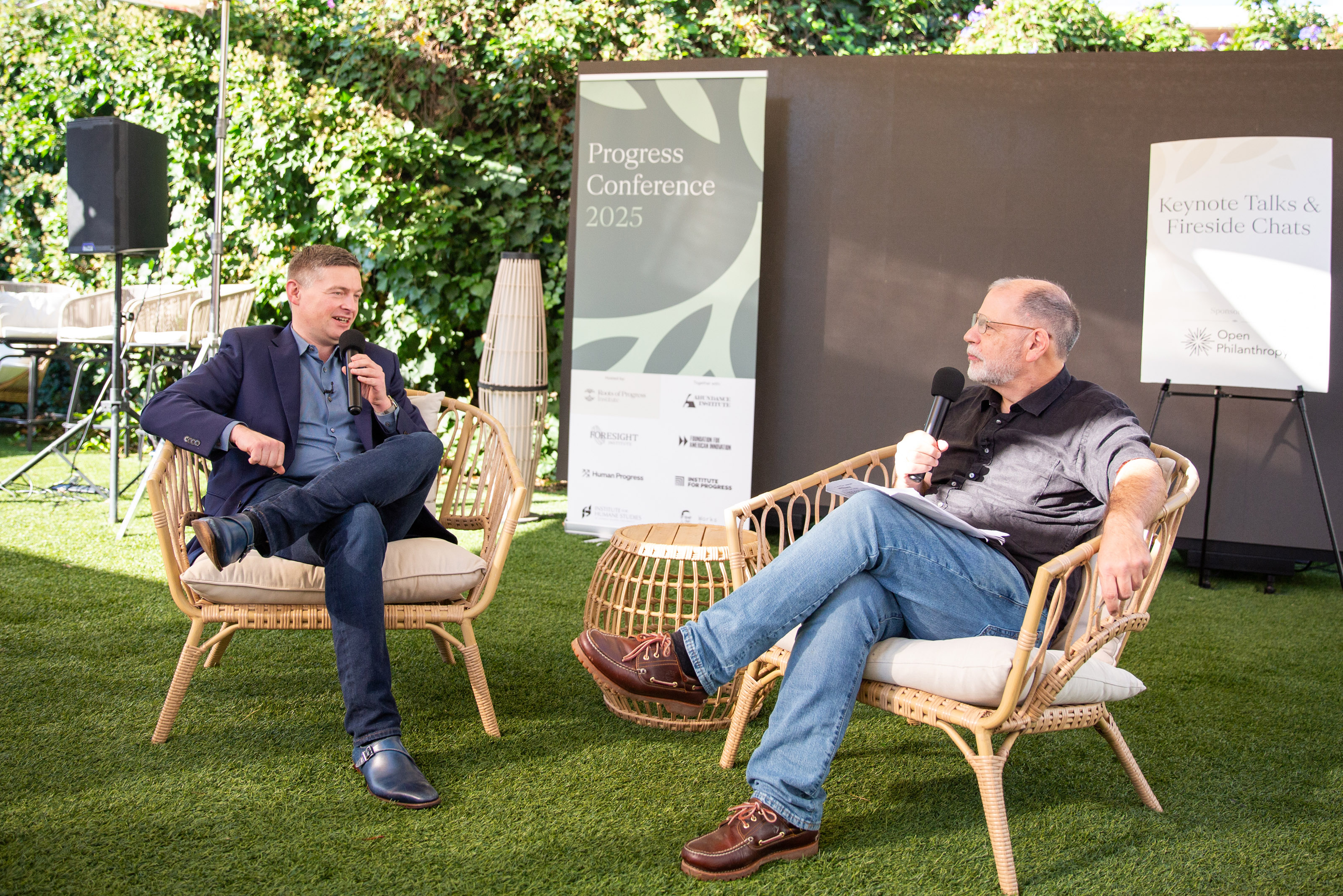 Two men sit on wicker chairs having a discussion with microphones at an outdoor event, with conference banners and greenery in the background.