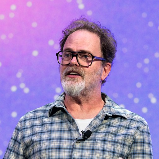 A bearded man wearing glasses and a plaid shirt speaks with a microphone clipped to his collar in front of a purple background with white dots.