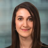 A woman with straight brown hair, wearing a dark blazer, smiles at the camera in front of a blurred background.
