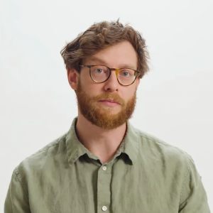 A man with curly brown hair, a beard, and glasses wears a light green collared shirt, standing against a plain white background.