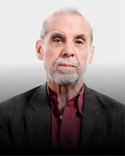 An older man with a light gray beard and slicked-back hair wears a dark jacket over a maroon shirt, looking directly at the camera against a plain light background.