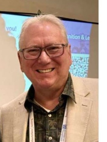 An older man wearing glasses, a patterned shirt, and a beige blazer smiles at an indoor event with a presentation screen in the background.