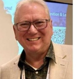 An older man wearing glasses, a patterned shirt, and a beige blazer smiles at an indoor event with a presentation screen in the background.