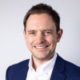 Man in a blue blazer and white shirt smiles at the camera against a plain light background.