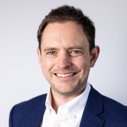 Man in a blue blazer and white shirt smiles at the camera against a plain light background.