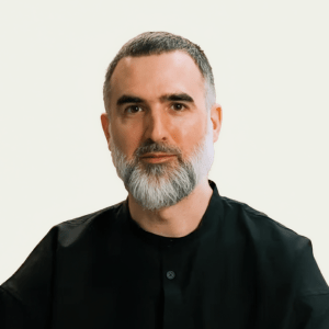 A man with short dark hair and a full gray beard wearing a black shirt sits against a plain light background, looking directly at the camera.