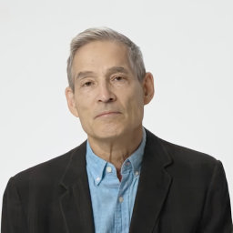 A middle-aged man with short gray hair wearing a light blue button-up shirt and a black blazer, standing against a plain white background.