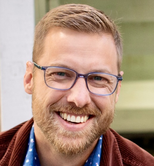 A man with short light brown hair, beard, and glasses is smiling. He is wearing a blue patterned shirt and a brown jacket. The background is out of focus.