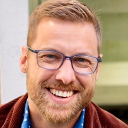 A man with short light brown hair, beard, and glasses is smiling. He is wearing a blue patterned shirt and a brown jacket. The background is out of focus.