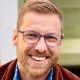 A man with short light brown hair, beard, and glasses is smiling. He is wearing a blue patterned shirt and a brown jacket. The background is out of focus.