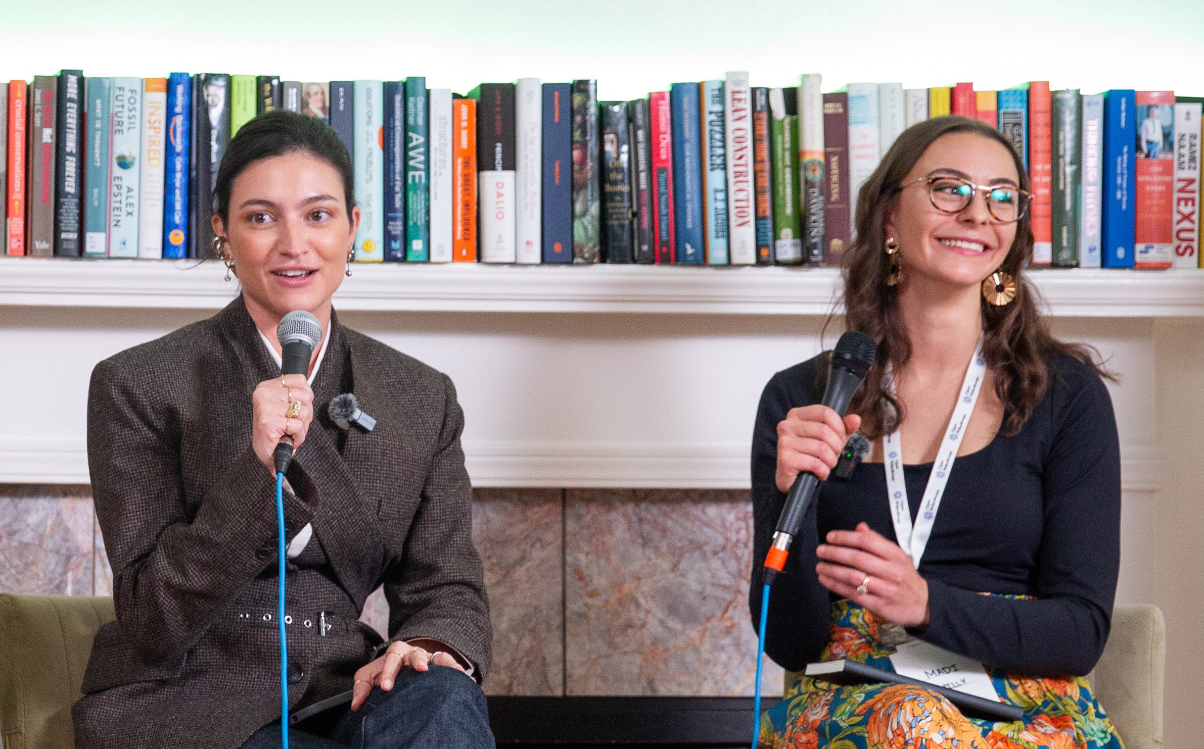 Two women sit in front of a bookshelf, each holding a microphone, engaged in conversation or discussion at an event.