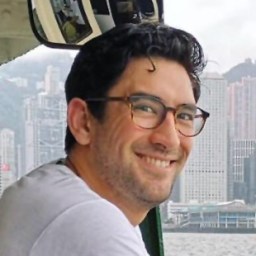 A man with dark hair and glasses smiles while leaning near a window, with city buildings and water visible in the background.