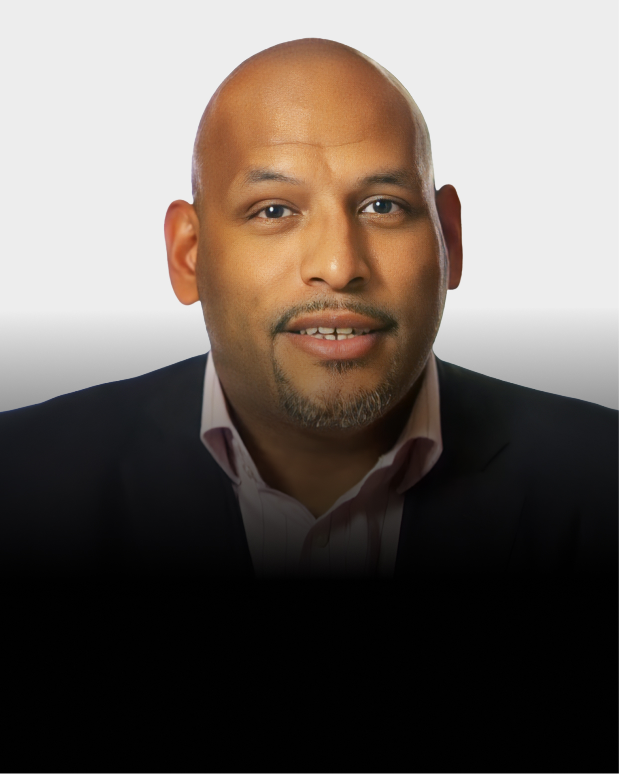 A man with a bald head, trimmed beard, and mustache, wearing a dark blazer and light-colored shirt, poses against a light background.