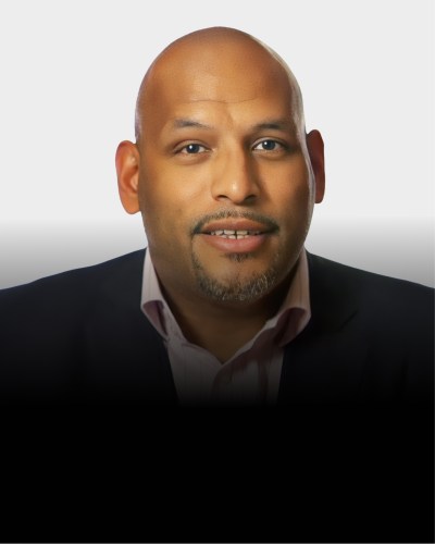 A man with a bald head, trimmed beard, and mustache, wearing a dark blazer and light-colored shirt, poses against a light background.