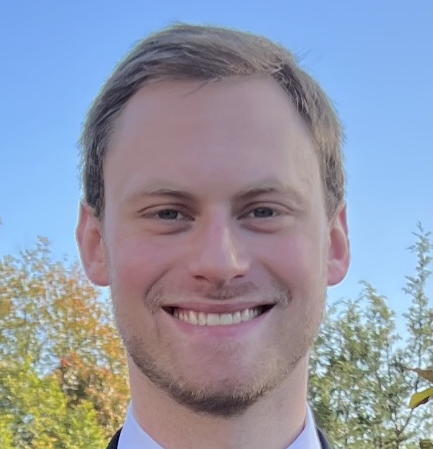 A young man with short light brown hair and light facial hair is smiling outdoors with trees and a clear blue sky in the background.