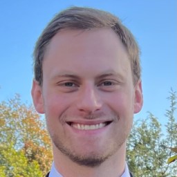 A young man with short light brown hair and light facial hair is smiling outdoors with trees and a clear blue sky in the background.