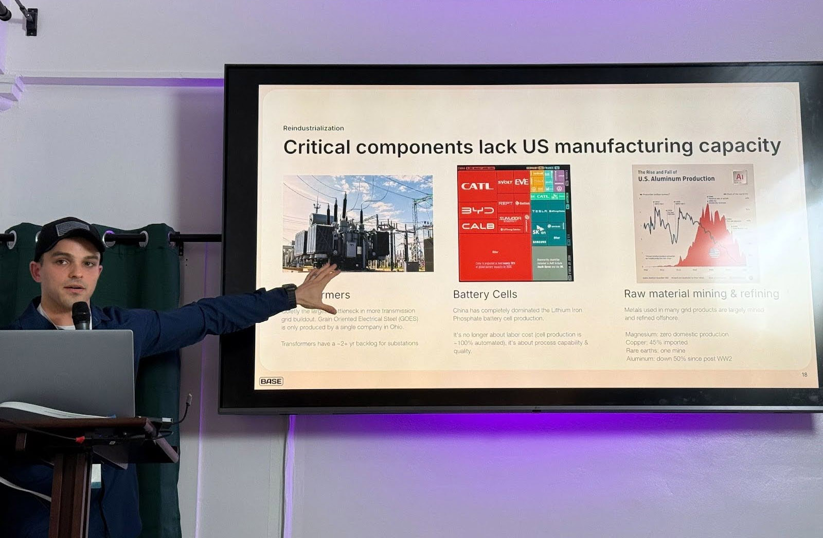 A man gives a presentation, pointing to a slide titled "Critical components lack US manufacturing capacity," featuring images and text on transformers, battery cells, and raw material mining.