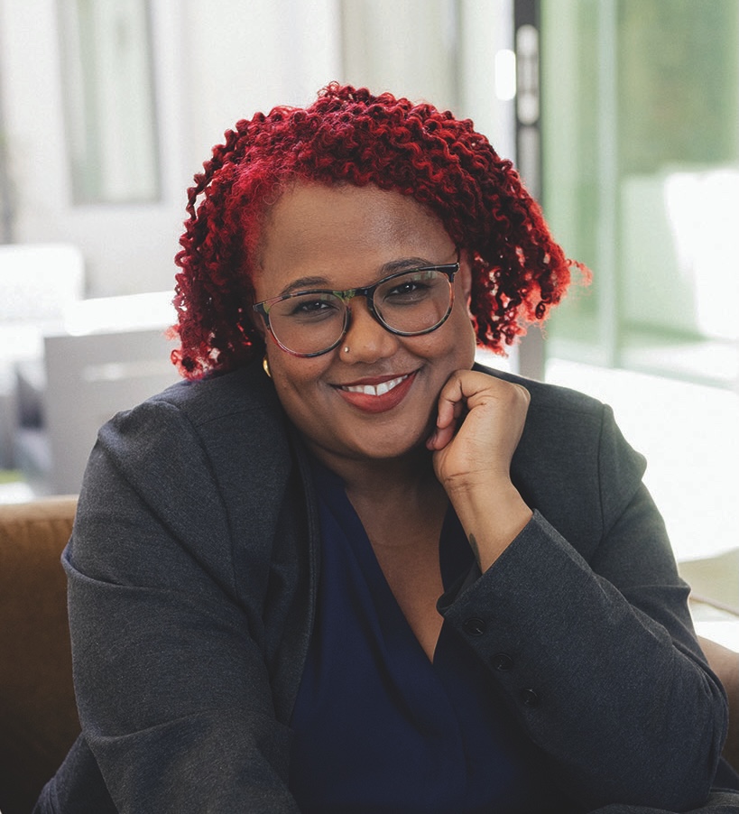 A person with red curly hair and glasses, wearing a dark blazer and navy shirt, smiles while resting their chin on their hand in a bright indoor setting.