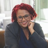 A person with red curly hair and glasses, wearing a dark blazer and navy shirt, smiles while resting their chin on their hand in a bright indoor setting.