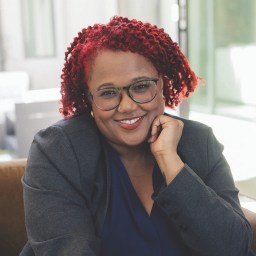 A person with red curly hair and glasses, wearing a dark blazer and navy shirt, smiles while resting their chin on their hand in a bright indoor setting.