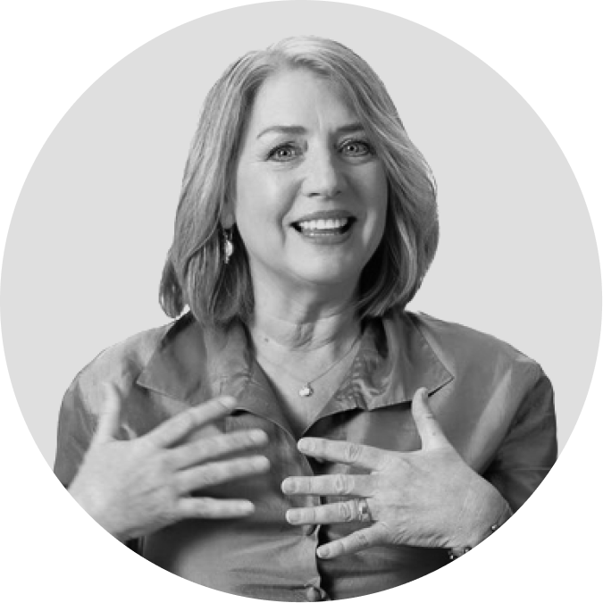 A woman with shoulder-length hair smiles and gestures with both hands on her chest while looking slightly to the side.