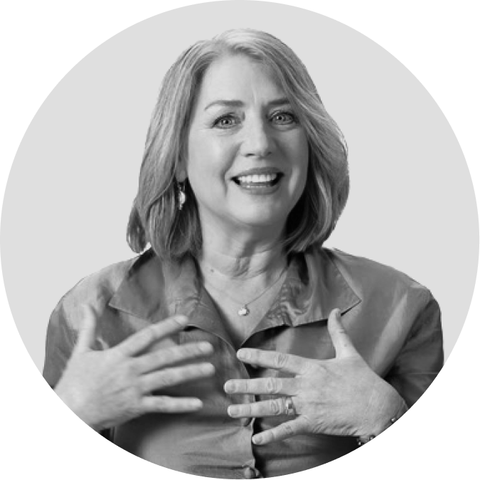 A woman with shoulder-length hair smiles and gestures with both hands on her chest while looking slightly to the side.