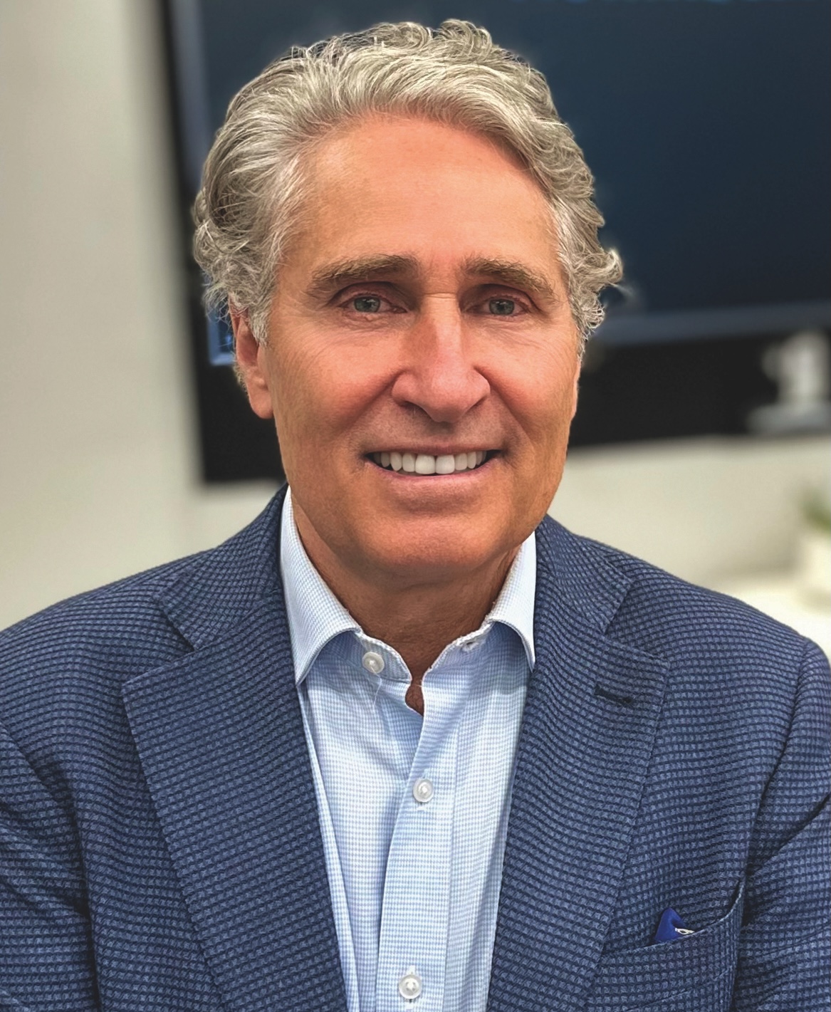 A man with gray, wavy hair wearing a blue textured blazer and light blue shirt, smiling in an indoor setting.