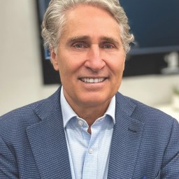 A man with gray, wavy hair wearing a blue textured blazer and light blue shirt, smiling in an indoor setting.