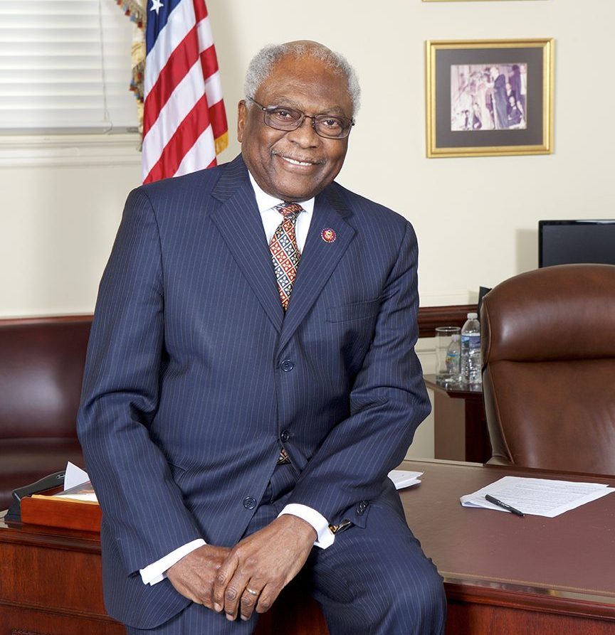 A man in a pinstripe suit sits on the edge of a wooden desk in an office with an American flag and framed photos on the wall.