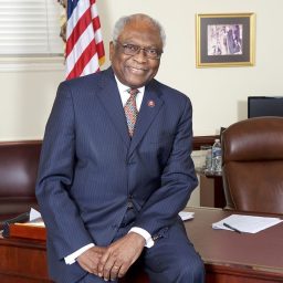 A man in a pinstripe suit sits on the edge of a wooden desk in an office with an American flag and framed photos on the wall.