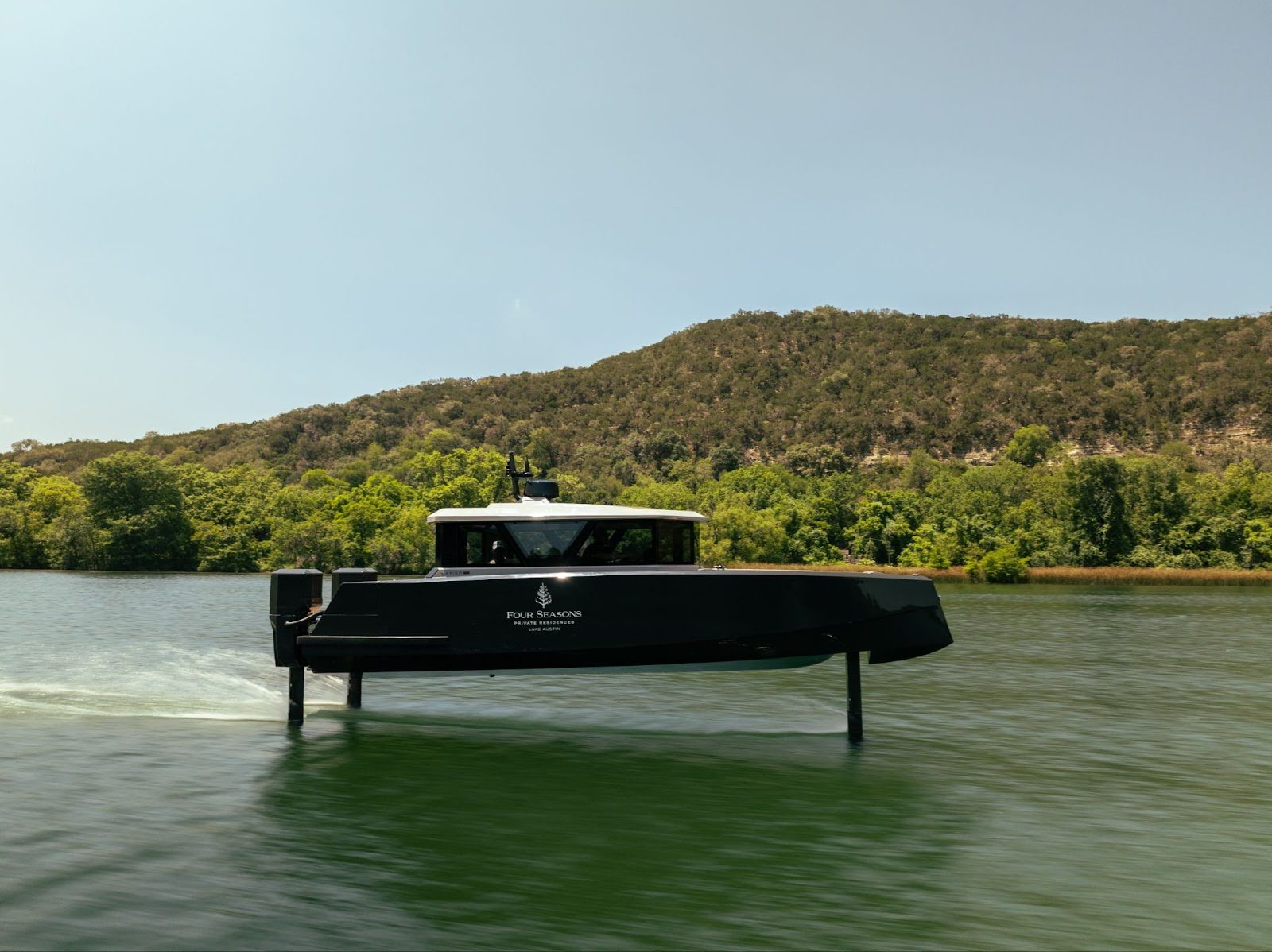 A black hydrofoil boat glides above the surface of a lake like one of the flying boats of legend, with green hills and trees in the background under a clear sky.