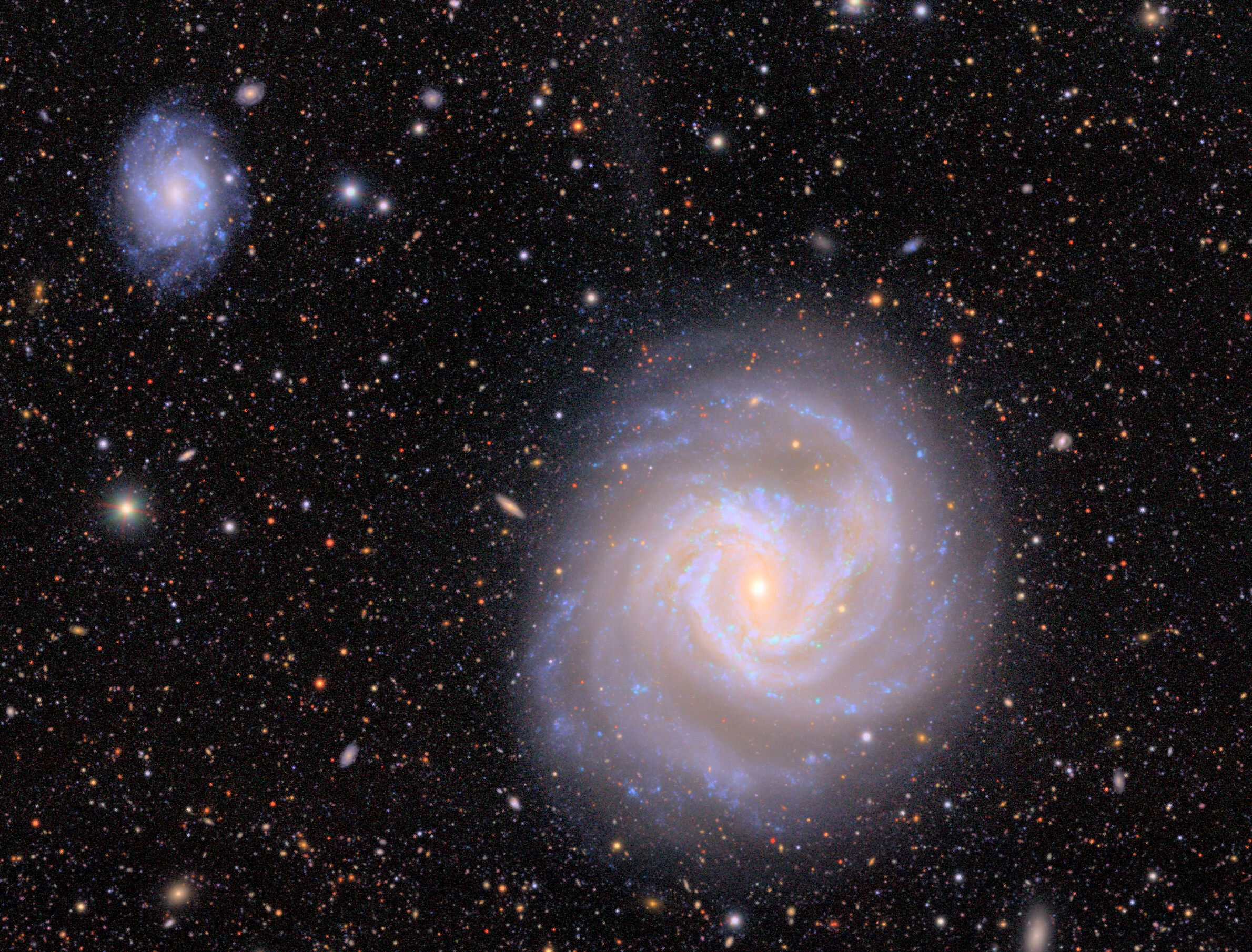 A bright spiral galaxy with a glowing center dominates the right, while a smaller, fainter galaxy in the upper left shows a stellar stream escaping its edge, all surrounded by numerous distant stars and galaxies.