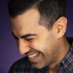 A man with short dark hair and stubble wearing a plaid shirt smiles and looks downward against a purple background.
