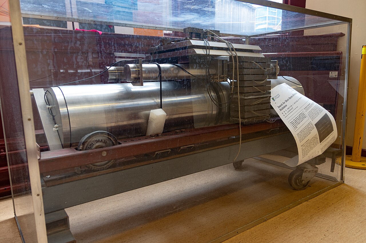 A large, metallic mechanical device&mdash;resembling those used to detect Weber bars and gravitational waves&mdash;is displayed in a glass case with an informational sheet attached. The industrial machine is set indoors on a tile floor.