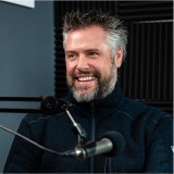 A man with gray hair and a beard is smiling while sitting in front of a microphone in a podcast studio.