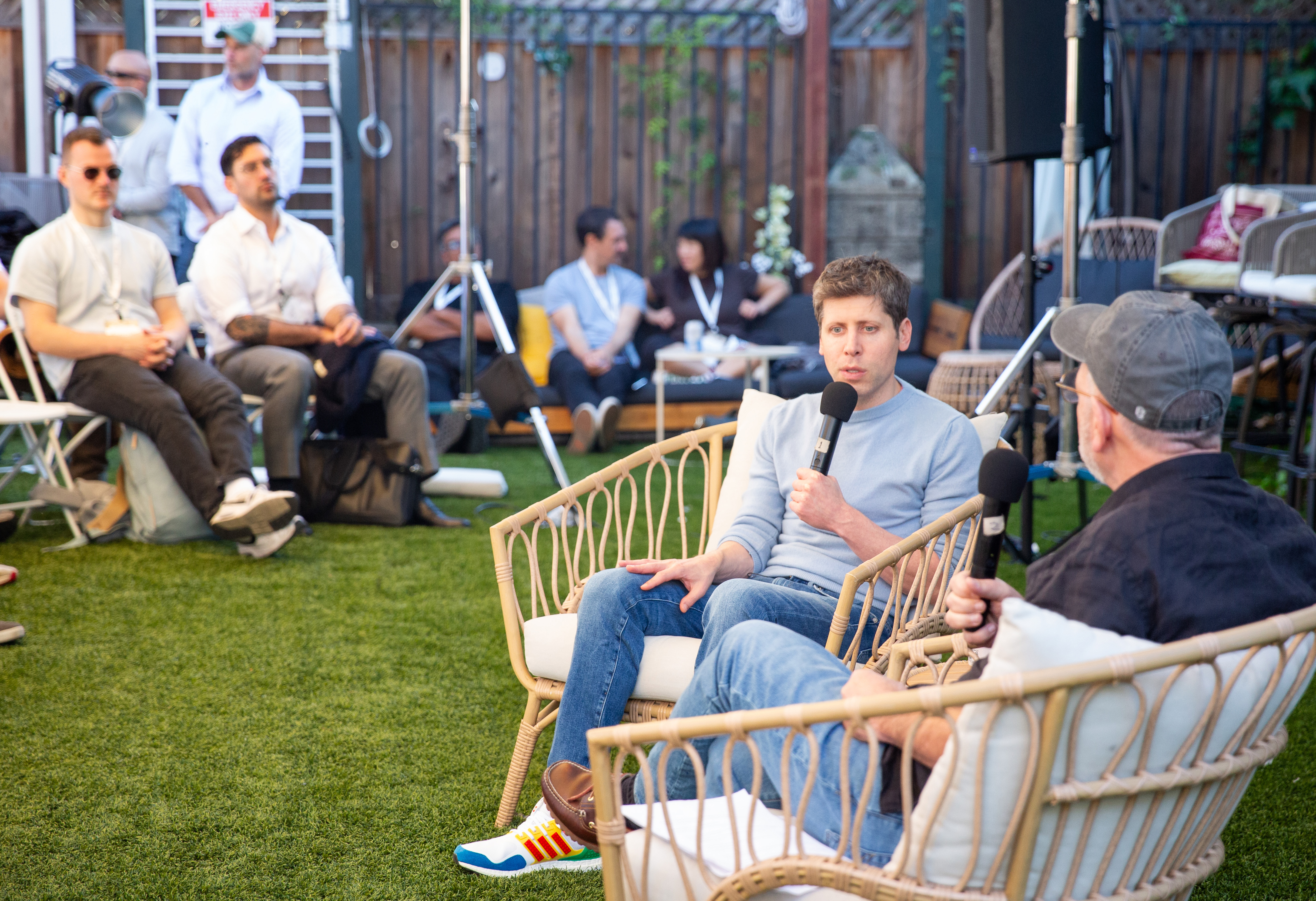 Two men sit on wicker chairs holding microphones, having a conversation outdoors while a small audience listens attentively in the background.