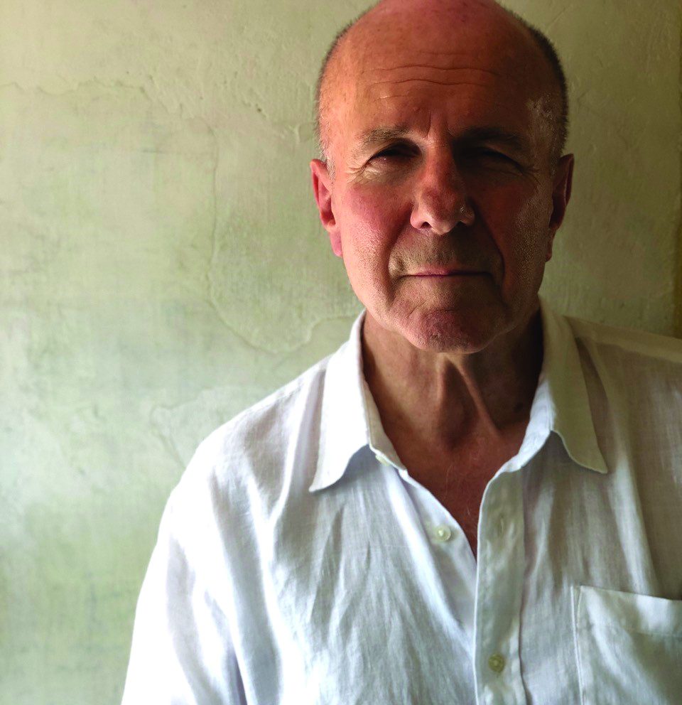 A man with a receding hairline wearing a white button-up shirt stands against a textured, light-colored wall.