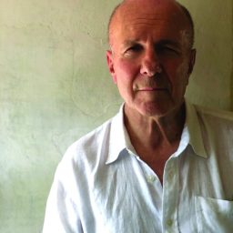 A man with a receding hairline wearing a white button-up shirt stands against a textured, light-colored wall.