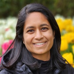 A person with long dark hair and a black jacket smiles outdoors in front of blooming yellow, white, and pink flowers.