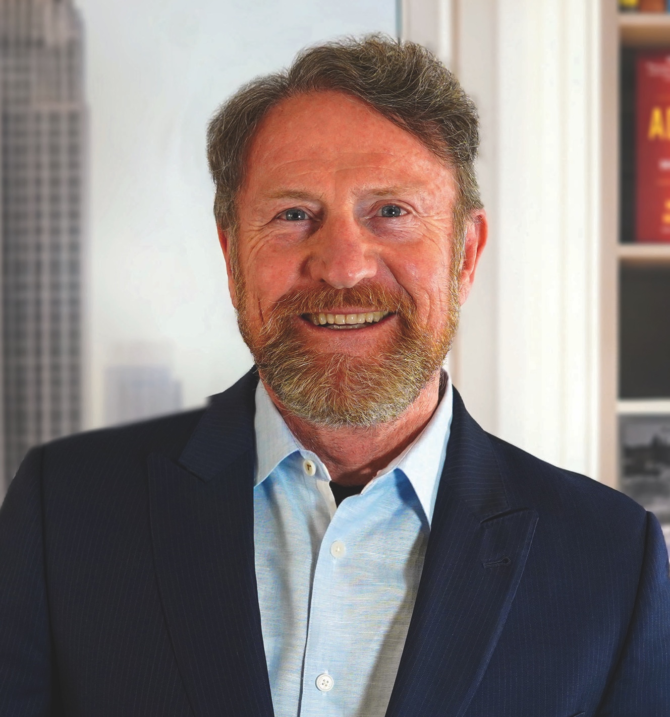 A middle-aged man with a beard and mustache, wearing a blue suit jacket and light blue shirt, standing indoors with bookshelves and a window in the background.